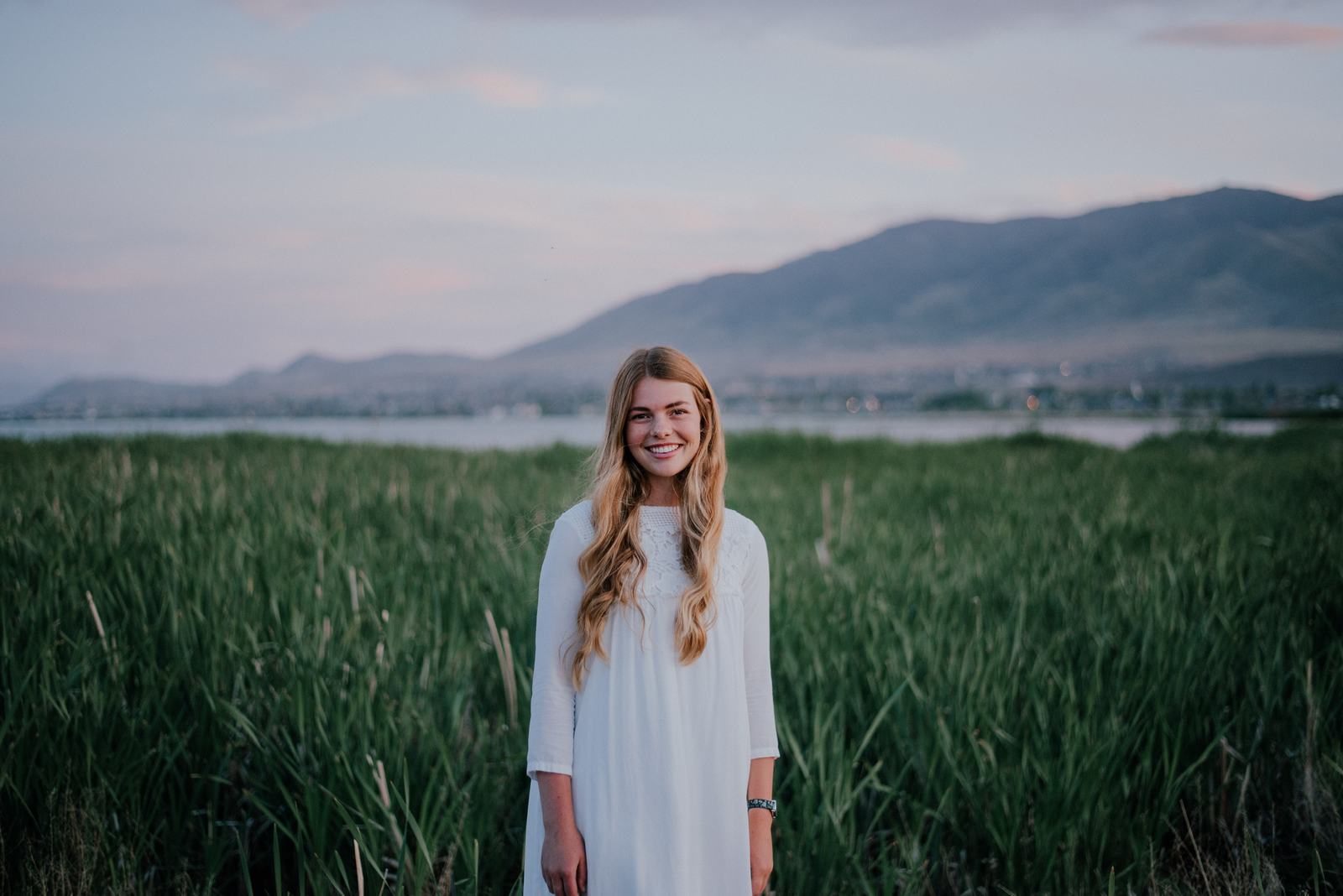 Allison smiling in a white dress, standing in a green field at dusk.