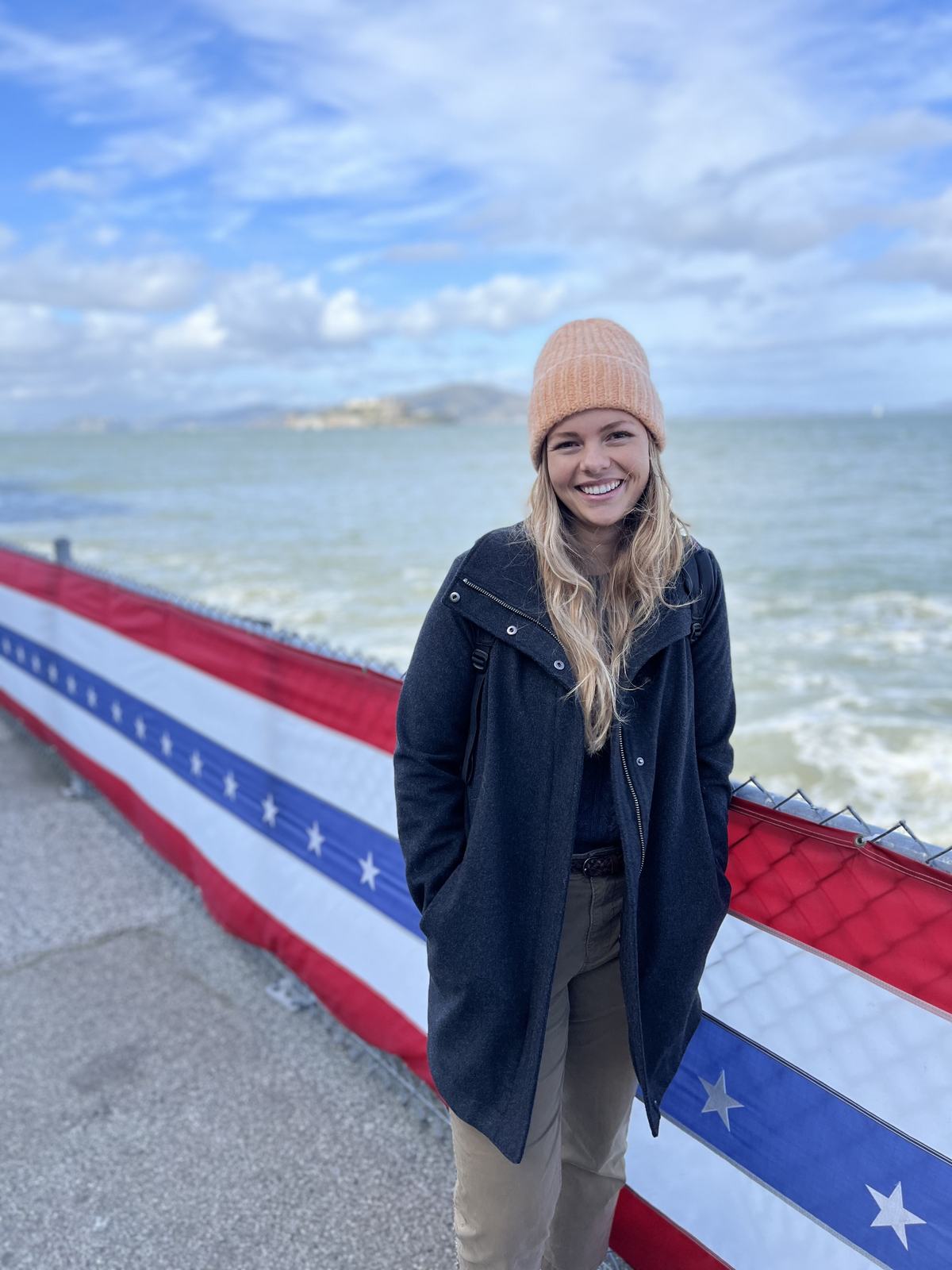 Allison in a peach beanie and dark coat, smiling by the water.